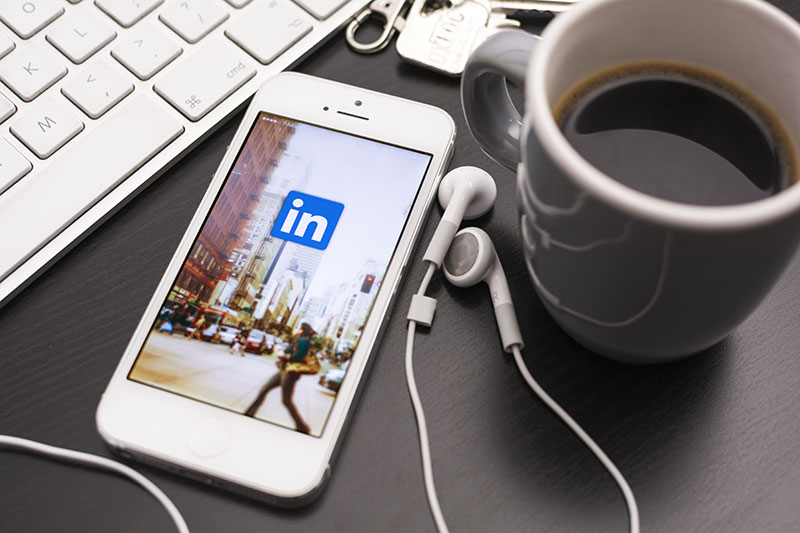 Linkedin app on a mobile phone with headphones, a coffee, and a keyboard.