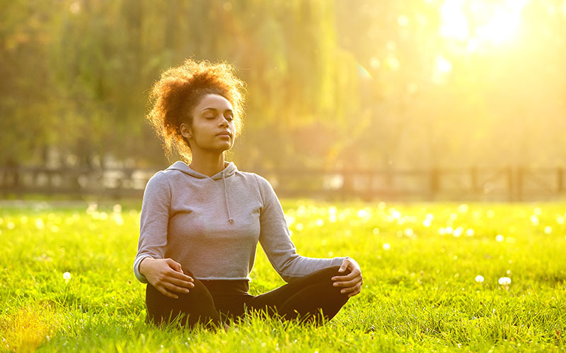 Female grad student meditating in a field with the sun shining on her.