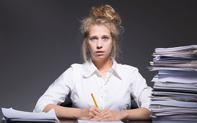 A frazzled female graduate student experiencing burnout sitting at a desk with piles of paper.