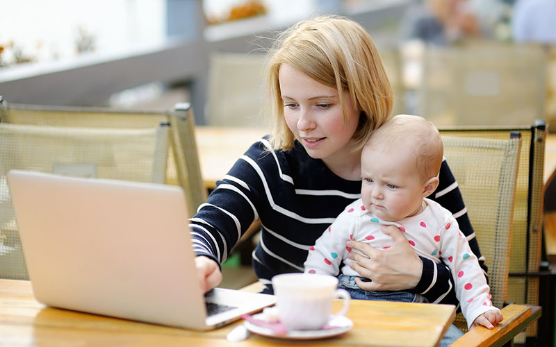 A female grad student who is a parent holding her baby while working on a laptop.