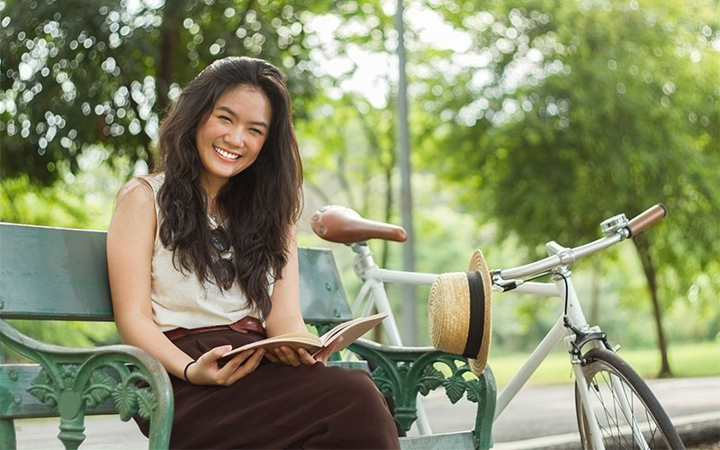 Female grad student sitting on a park bench beside her bicycle smiling and reading a book.