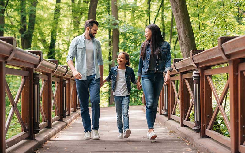 A mother and father walking on an outdoor boardwalk in the woods with their little girl.